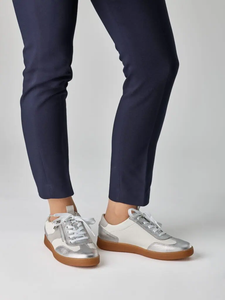 Damskor i silver och vit med gummisula, nära benen på en person i mörkblå byxor.